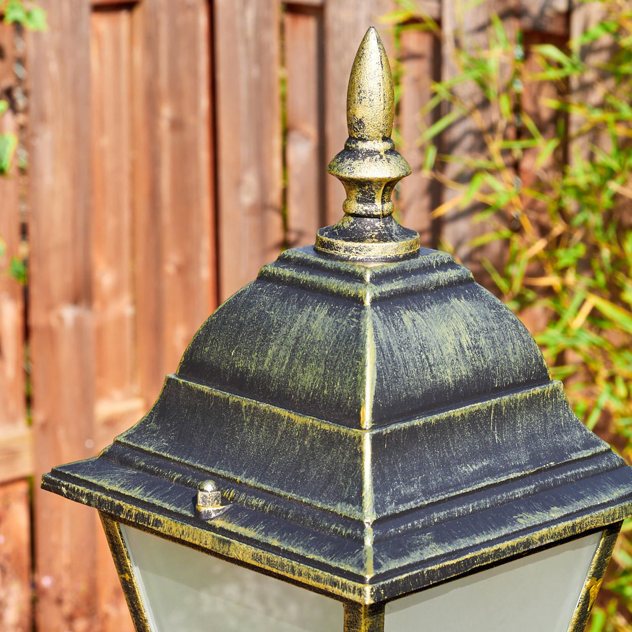 Nahaufnahme einer schwarzen und goldenen Außenlampe. Ein Holzzaun und grüner Bambus sind im verschwommenen Hintergrund.