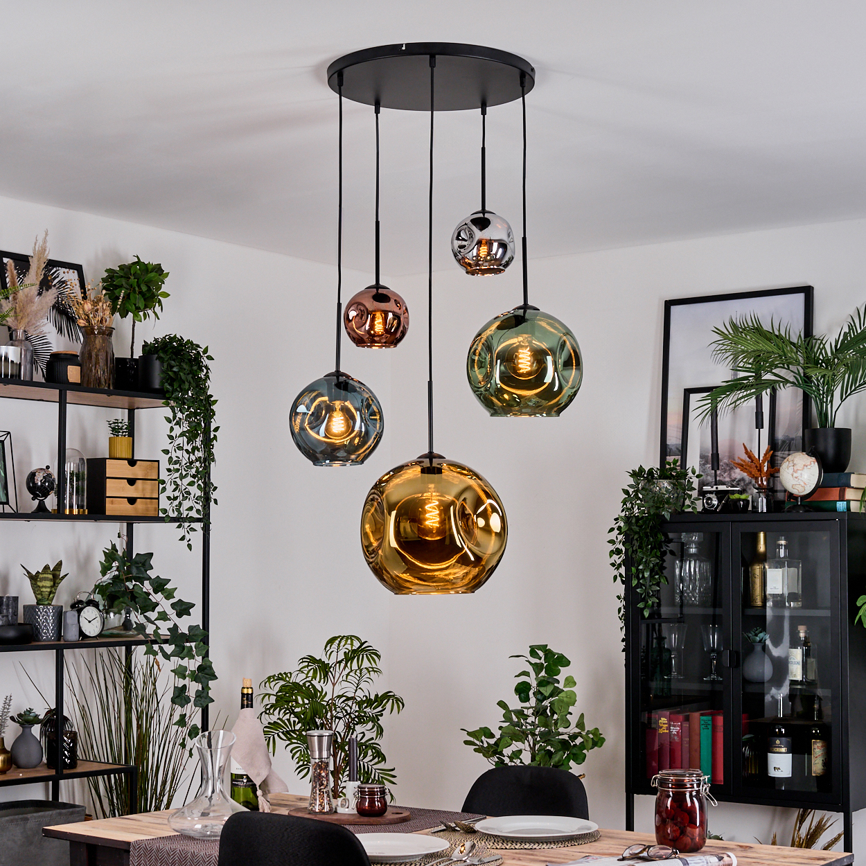 Esszimmer mit hängenden Glaslampen. Pflanzen, Regale, ein Tisch mit Gedeck und ein Schrank sind im Raum zu sehen.