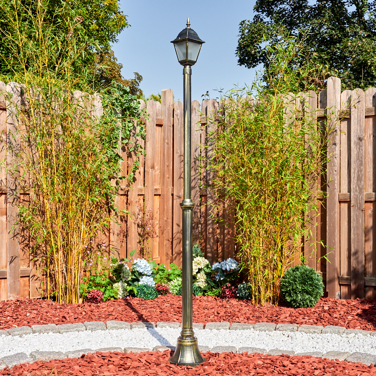 Eine Gartenszene mit Laterne, Bambus, Blumen und Holzzaun. Der Boden hat roten Mulch mit einem weißen Steinweg.