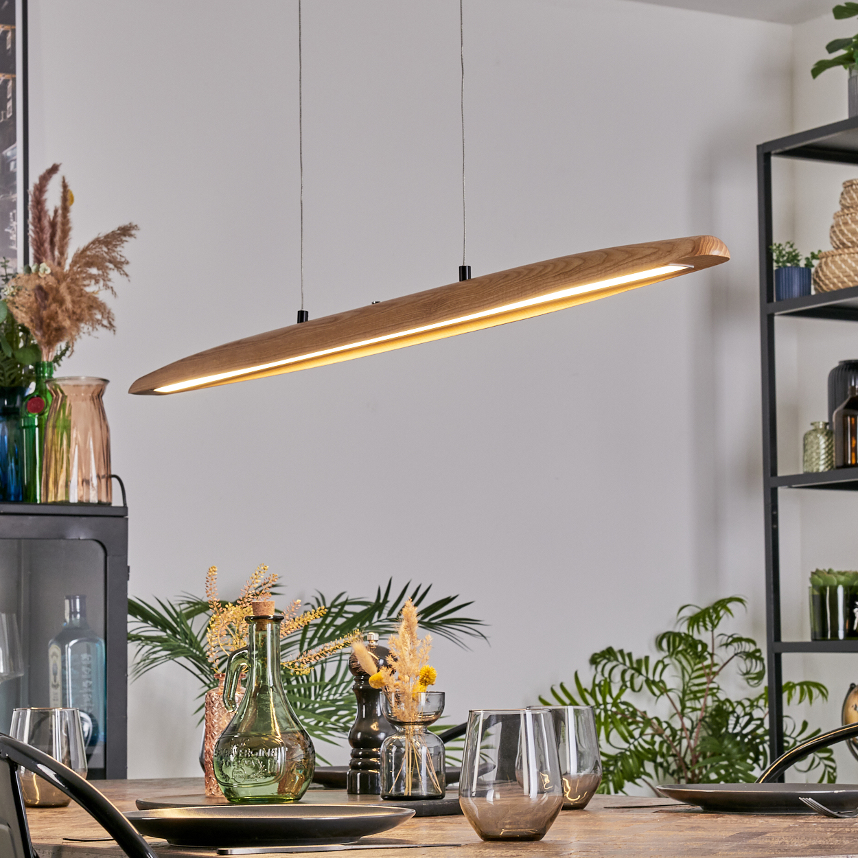 Holzlampe hängt über Tisch mit Gläsern, Pflanzen und Vasen. Regale mit Dekoration im Hintergrund. Helle Beleuchtung.