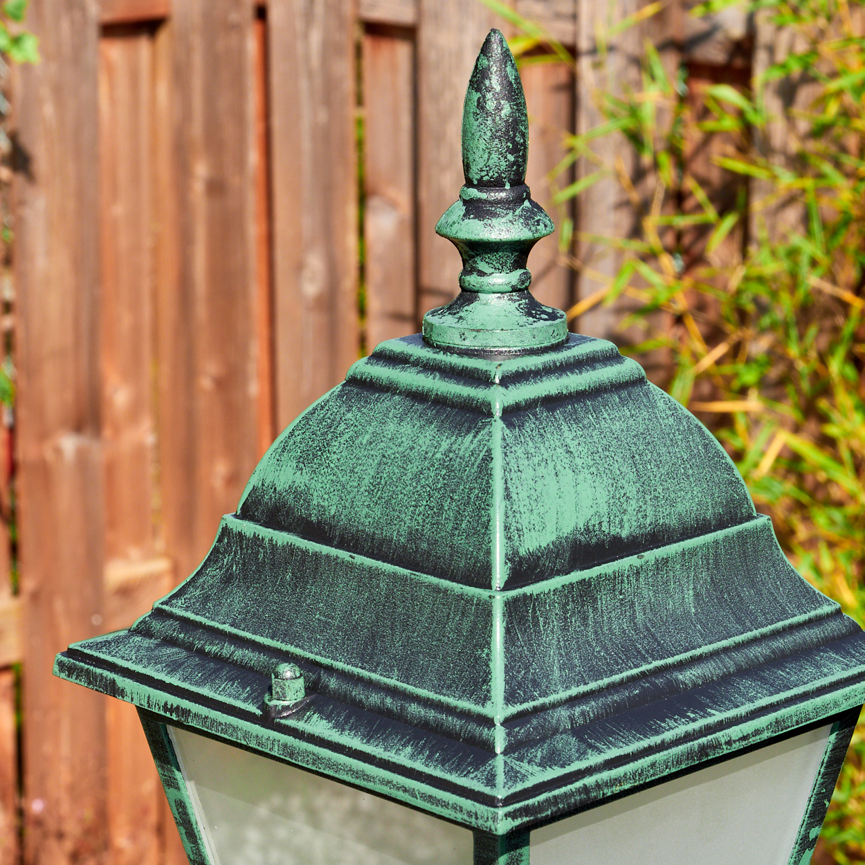 Eine grüne und schwarze Außenlampe. Der Hintergrund zeigt einen Holzzaun und grüne Pflanzen. Die Lampe ist im Fokus.