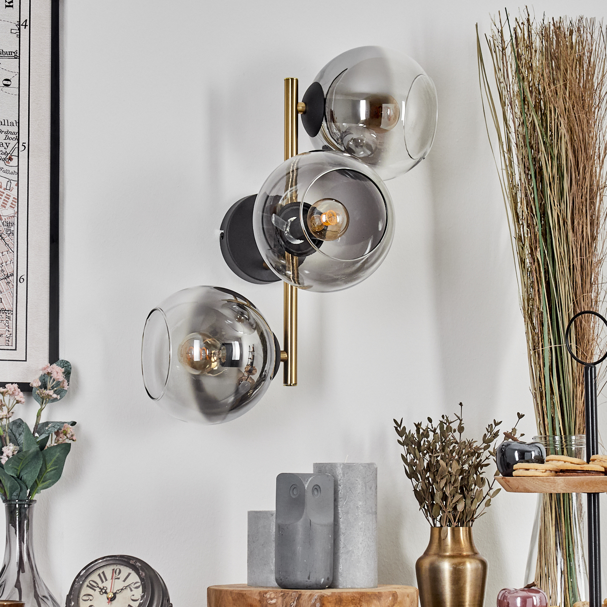 Eine moderne Wandlampe mit drei Glaskugeln. Im Vordergrund befinden sich verschiedene Dekorationsartikel, darunter eine Uhr und Blumen.