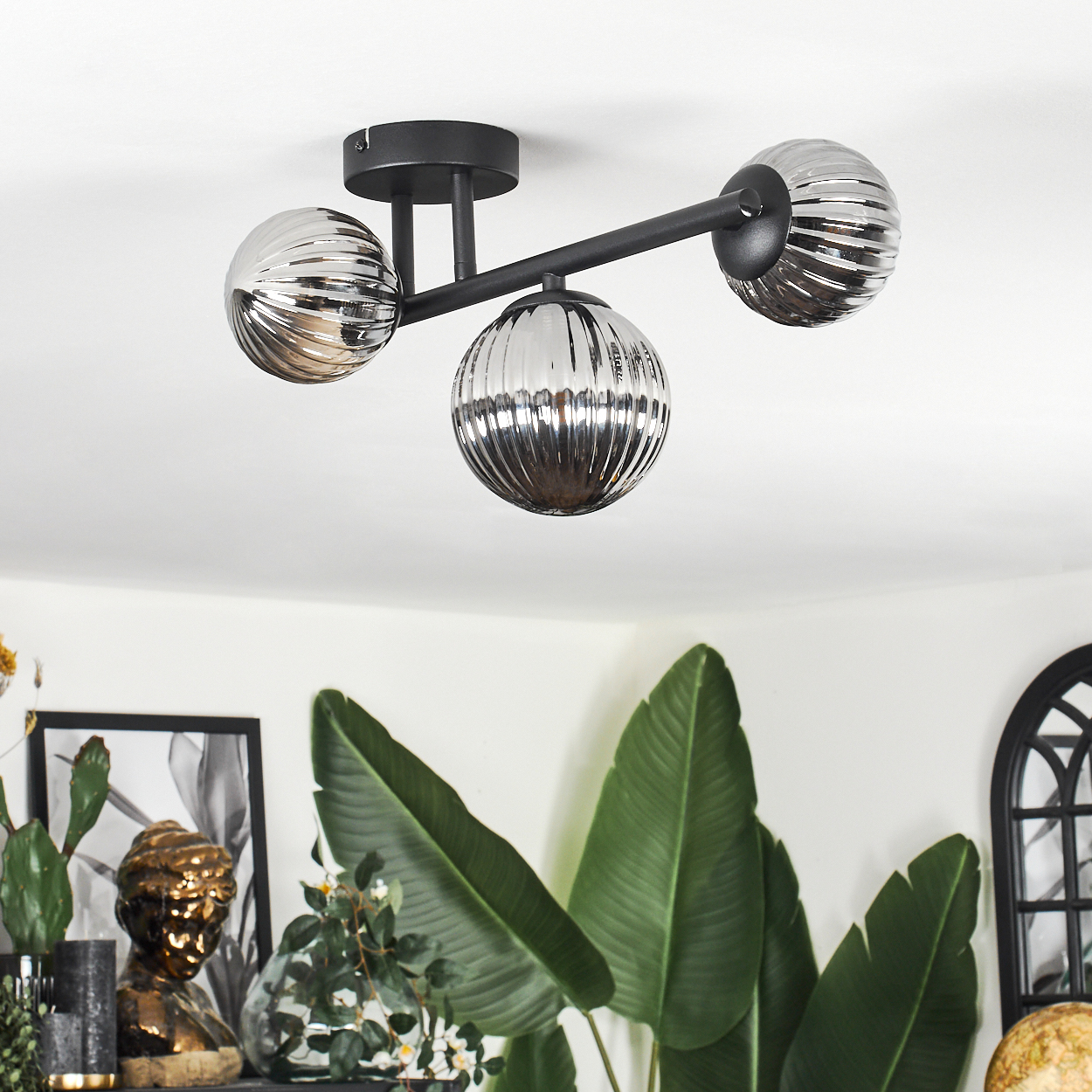 Eine moderne Deckenlampe mit drei kugelförmigen Schirmen. Darunter sind große grüne Blätter und Dekorationen zu sehen.
