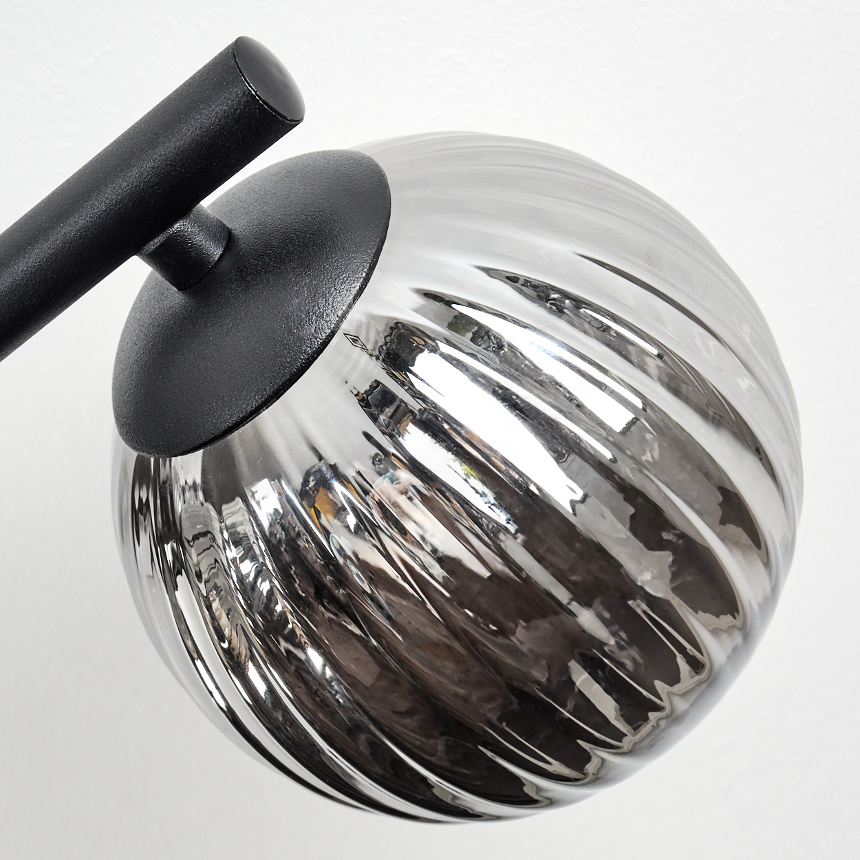 Nahaufnahme einer modernen Lampe. Sie hat einen schwarzen Arm und eine silberne, gerippte Glaskugel. Der Hintergrund ist eine weiße Wand.