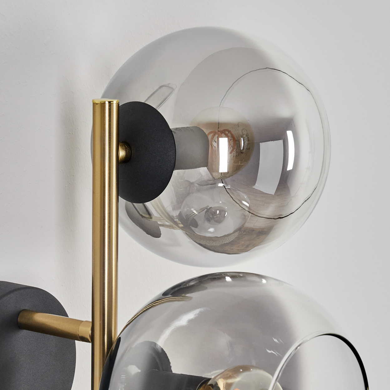 Nahaufnahme einer Wandlampe. Sie hat zwei Glaskugeln und goldfarbene Akzente. Der Hintergrund ist eine weiße Wand.