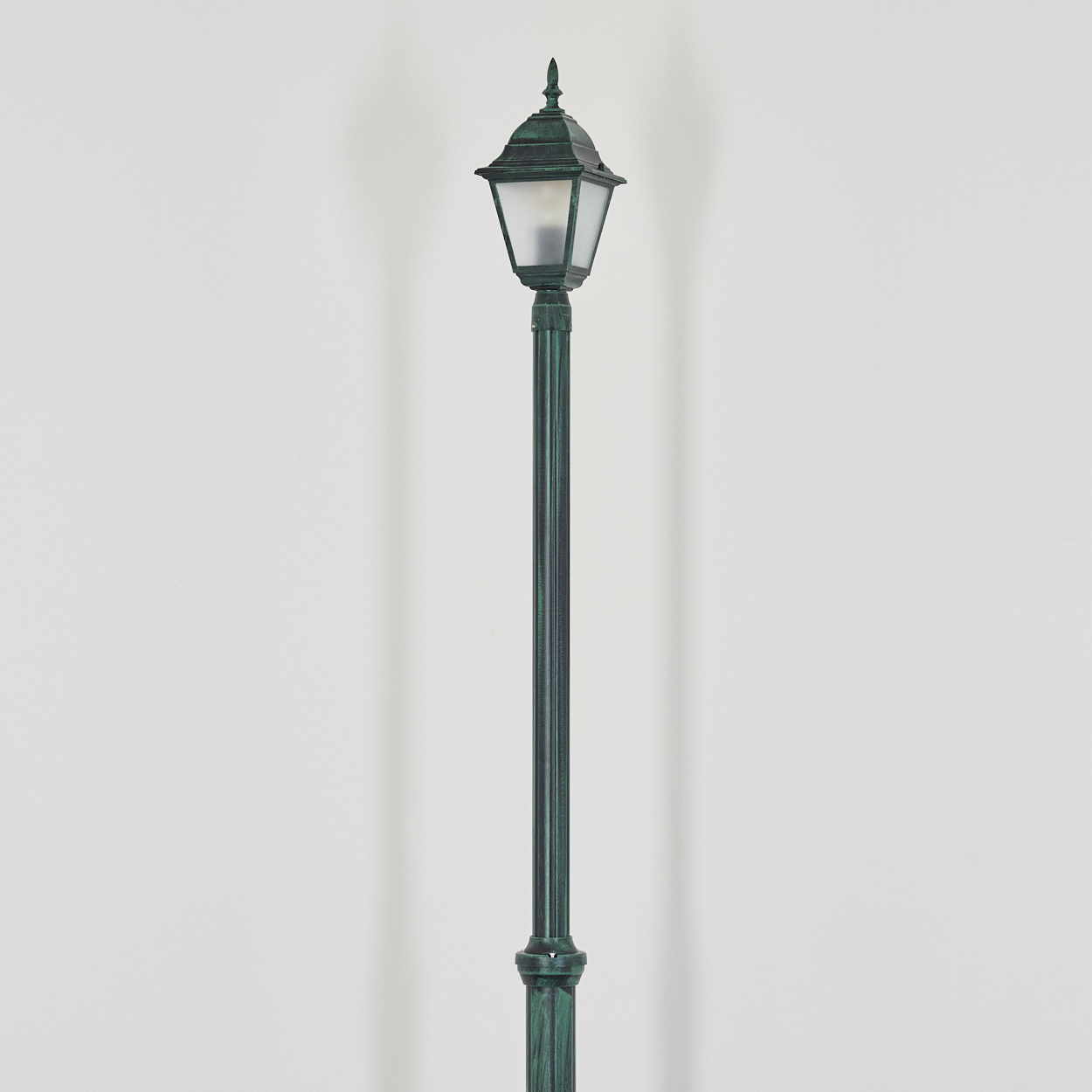 Eine verzierte grüne Straßenlaterne steht mittig vor einer weißen Wand. Die Lampe hat eine spitze Spitze und einen matten Glasschirm.