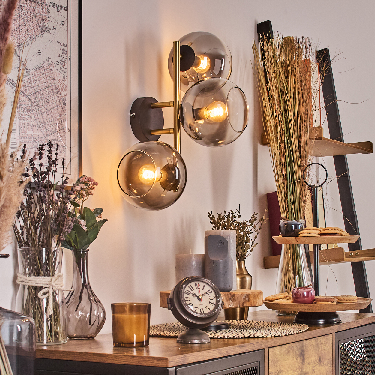 Eine dekorative Wandlampe mit drei kugelförmigen Schirmen. Verschiedene Dekorationen, darunter Vasen, Kerzen und eine Uhr, befinden sich auf Holz.