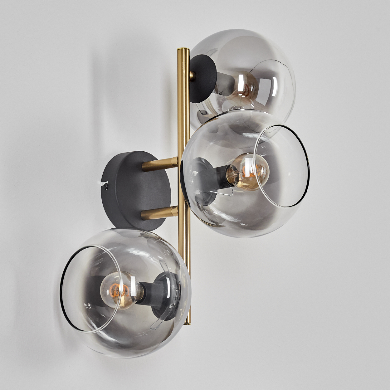 Eine moderne Wandlampe mit drei Glaskugelschirmen. Sie hat messingfarbene Arme und einen schwarzen Sockel, montiert an einer weißen Wand.