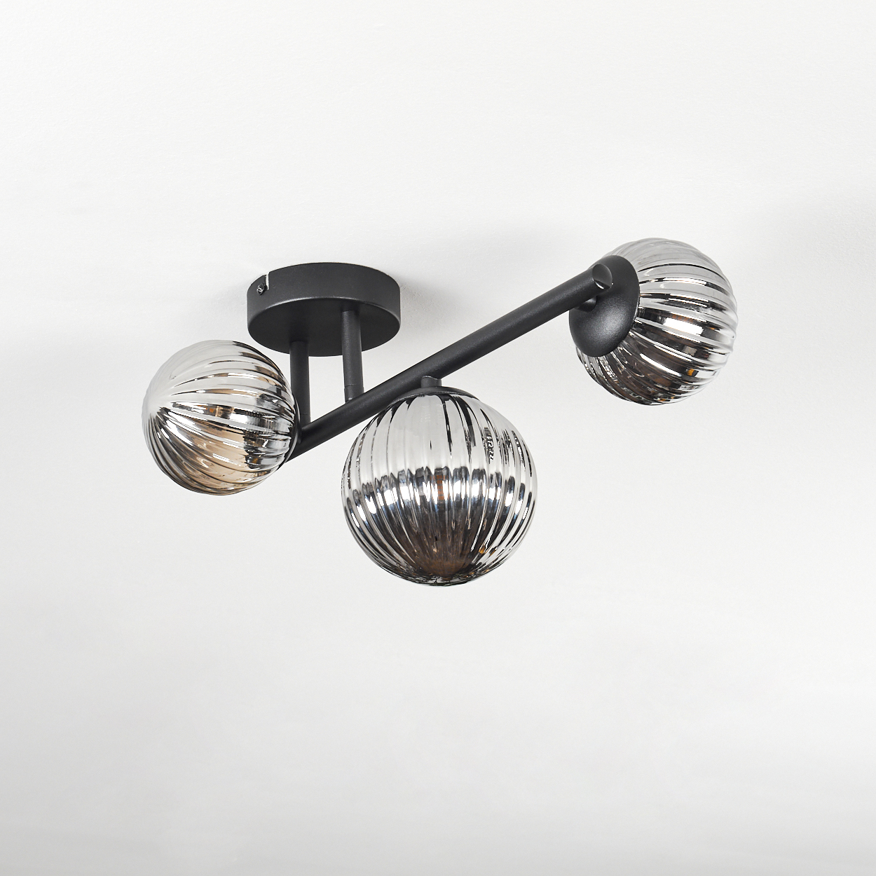 Moderne Deckenleuchte mit drei kugelförmigen, gerippten Glasschirmen. Sie ist an einem schwarzen Metallrahmen an einer weißen Decke befestigt.
