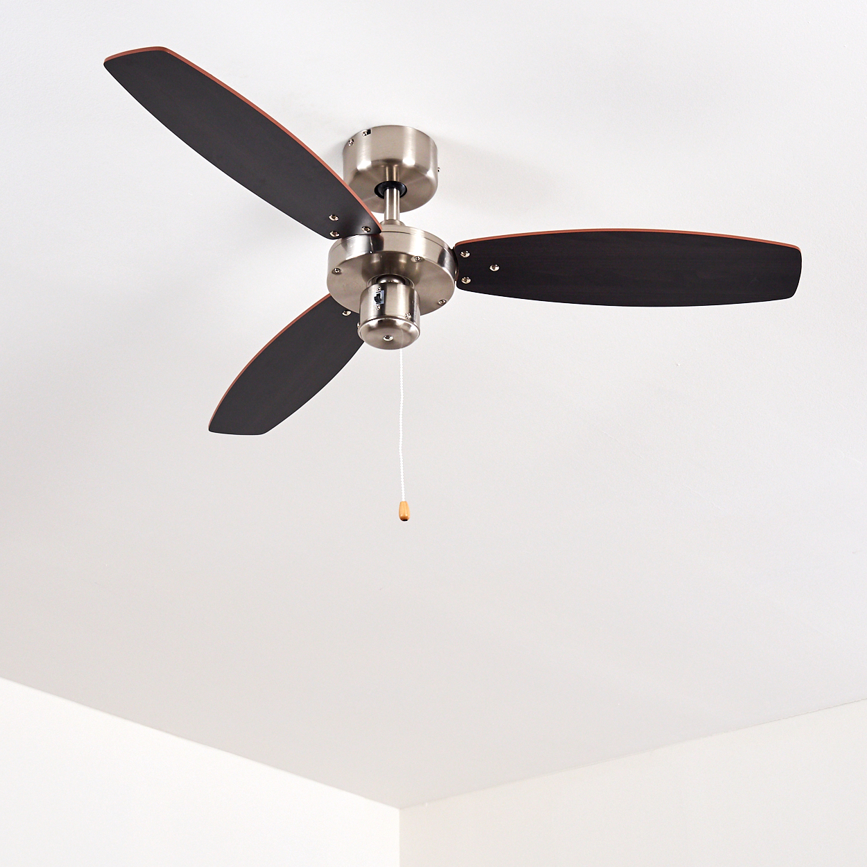 Ein Deckenventilator mit drei dunkelbraunen Blättern und einem silbernen Motor hängt an einer weißen Decke. Eine Zugschnur hängt herunter.