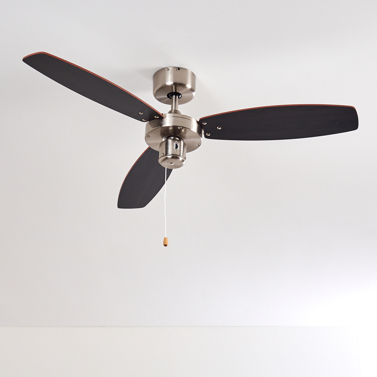 Ein Deckenventilator mit drei dunklen Flügeln und einem silbernen Motor hängt von einer weißen Decke. Ein Zugband hängt herunter.