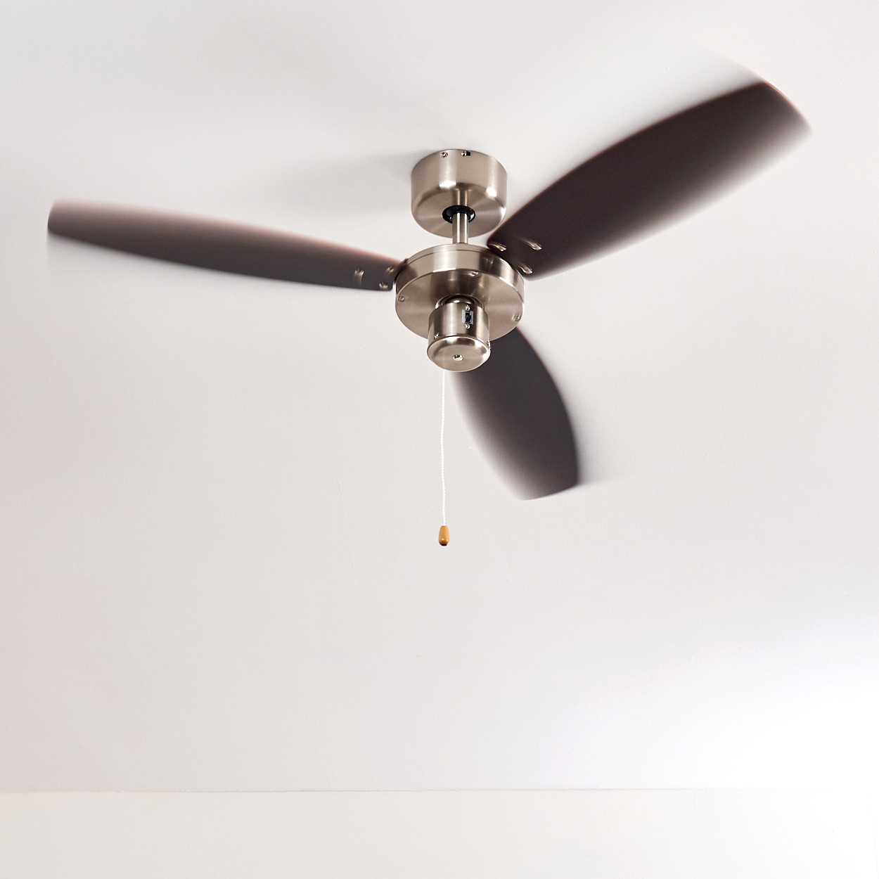 Ein Deckenventilator mit drei Flügeln dreht sich. Die Flügel sind dunkel und der Motor ist silberfarben. Ein Zugband hängt herunter.