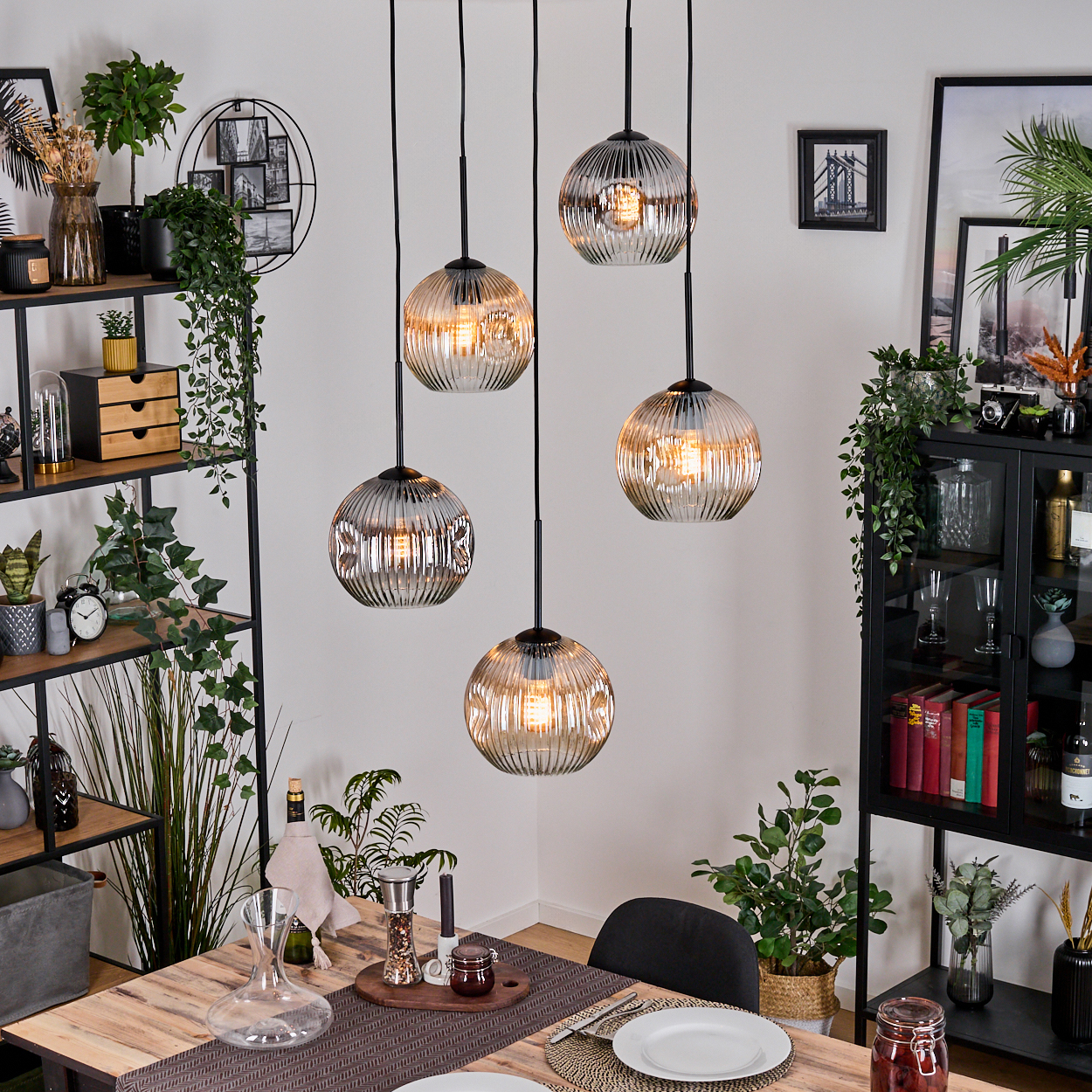 Esszimmer mit hängenden Kugelleuchten. Tisch mit Tellern, Pflanzen und Regalen mit Dekor. Schwarze Metallregale und Schränke.