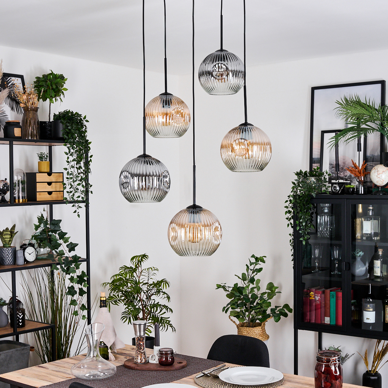 Esszimmer mit Hängelampen. Ein Tisch ist gedeckt, Pflanzen und Deko. Regale und ein Schrank zeigen weitere dekorative Artikel.