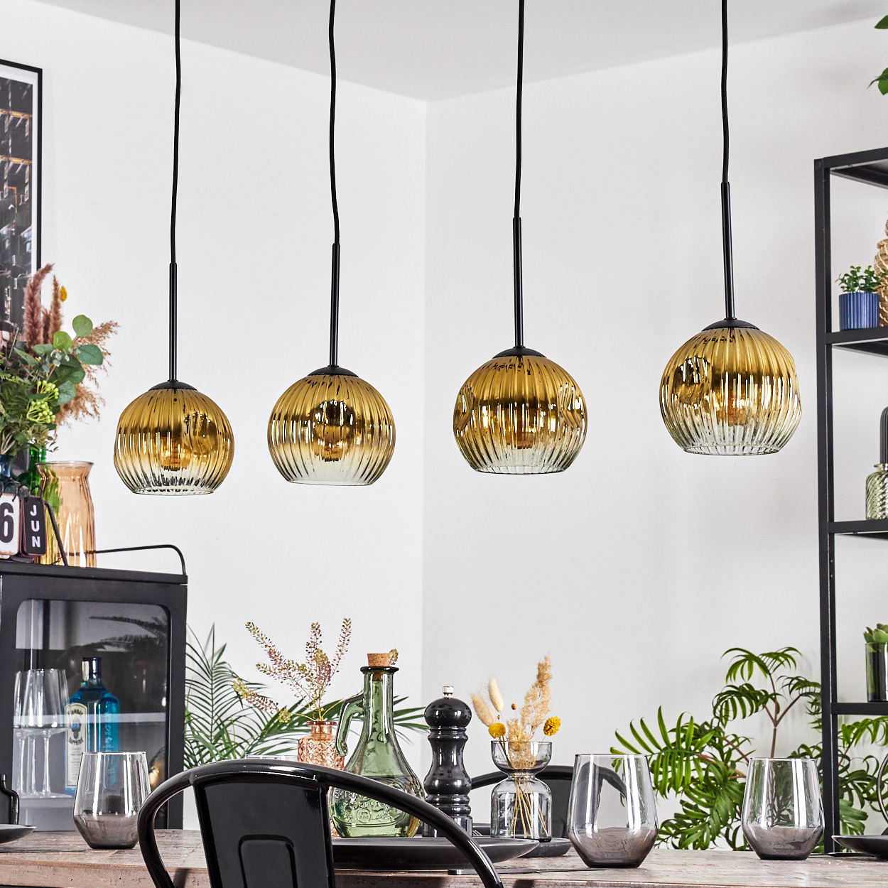 Vier goldene Pendelleuchten hängen über einem Esstisch mit Gläsern und Dekorationen. Im Hintergrund befindet sich ein schwarzes Regal.