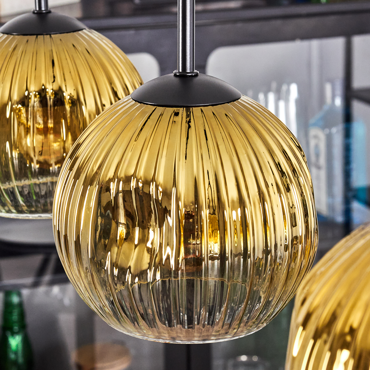 Drei goldene, gerippte Glas-Pendelleuchten hängen. Schwarze Armaturen und Reflexionen sind sichtbar. Im Hintergrund sind Flaschen und Regale zu sehen.