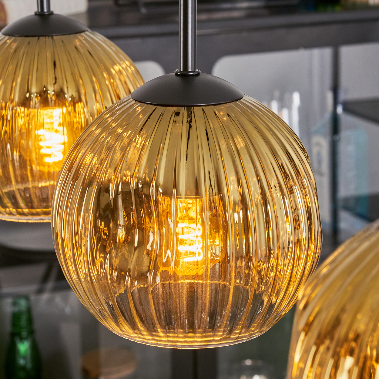 Drei goldene, geriffelte Glas-Pendelleuchten mit freiliegenden Glühbirnen, die an einer dunklen Halterung hängen. Der Hintergrund ist verschwommen.
