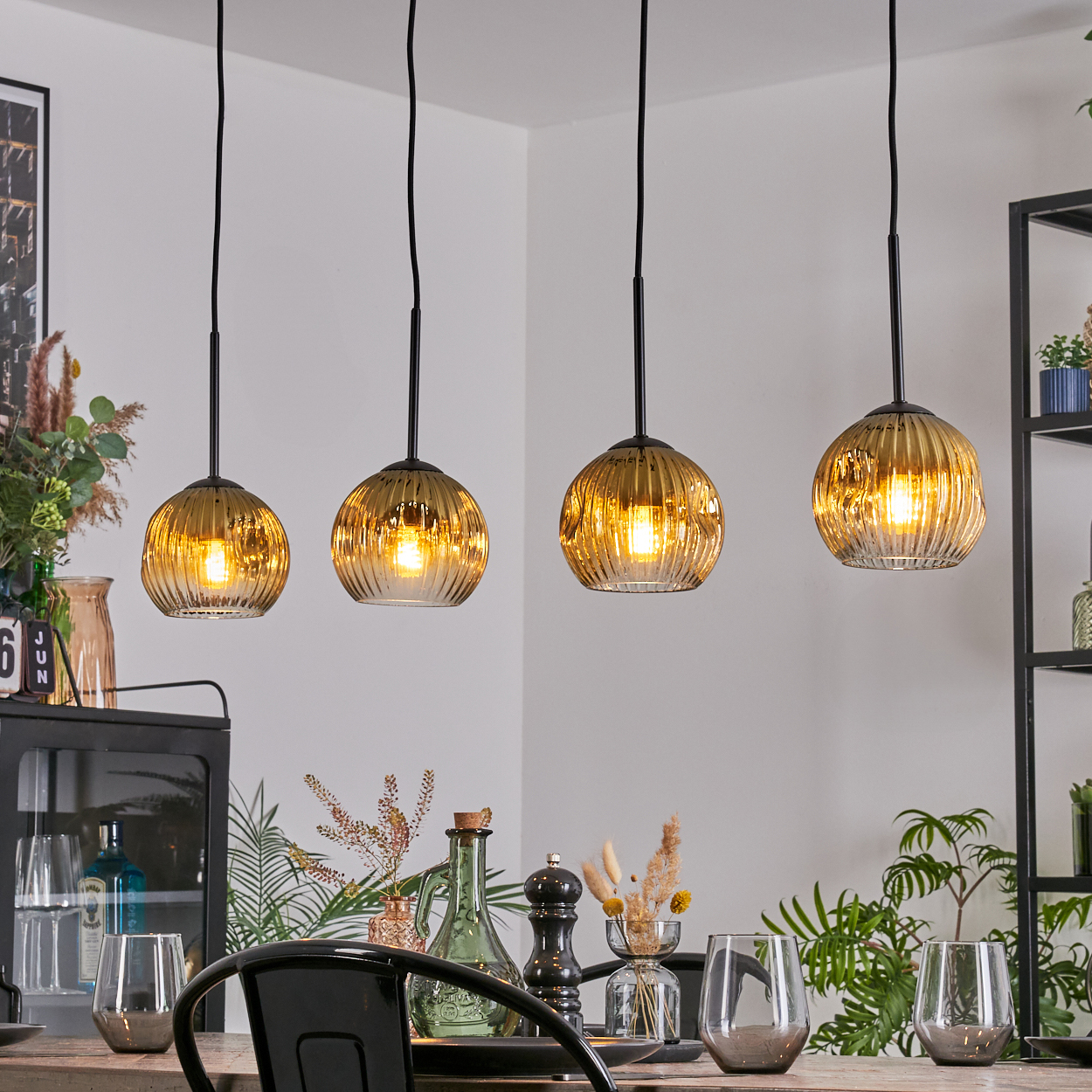Vier bernsteinfarbene Glas-Pendelleuchten hängen über einem Esstisch. Tisch mit Gläsern und Dekor. Pflanzen und Regale im Hintergrund.