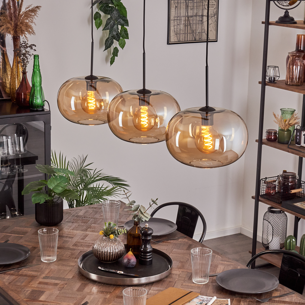 Esszimmer-Szene mit drei bernsteinfarbenen Pendelleuchten über einem Holztisch, der zum Abendessen gedeckt ist. Pflanzen, Dekor und Regale sind im Hintergrund.