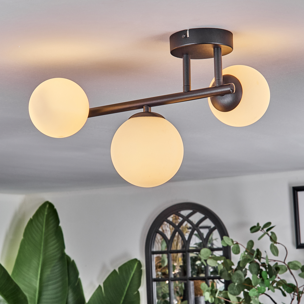 Eine moderne Deckenleuchte mit drei kugelförmigen Lampen. Das Licht ist braun und die Glühbirnen sind weiß. Im Hintergrund grüne Pflanzen und ein Fenster.