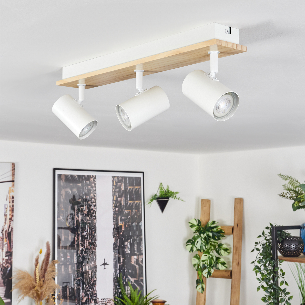 Drei weiße Strahler an einem Holzbalken an einer weißen Decke. Kunstwerke und Pflanzen sind im Hintergrund sichtbar.