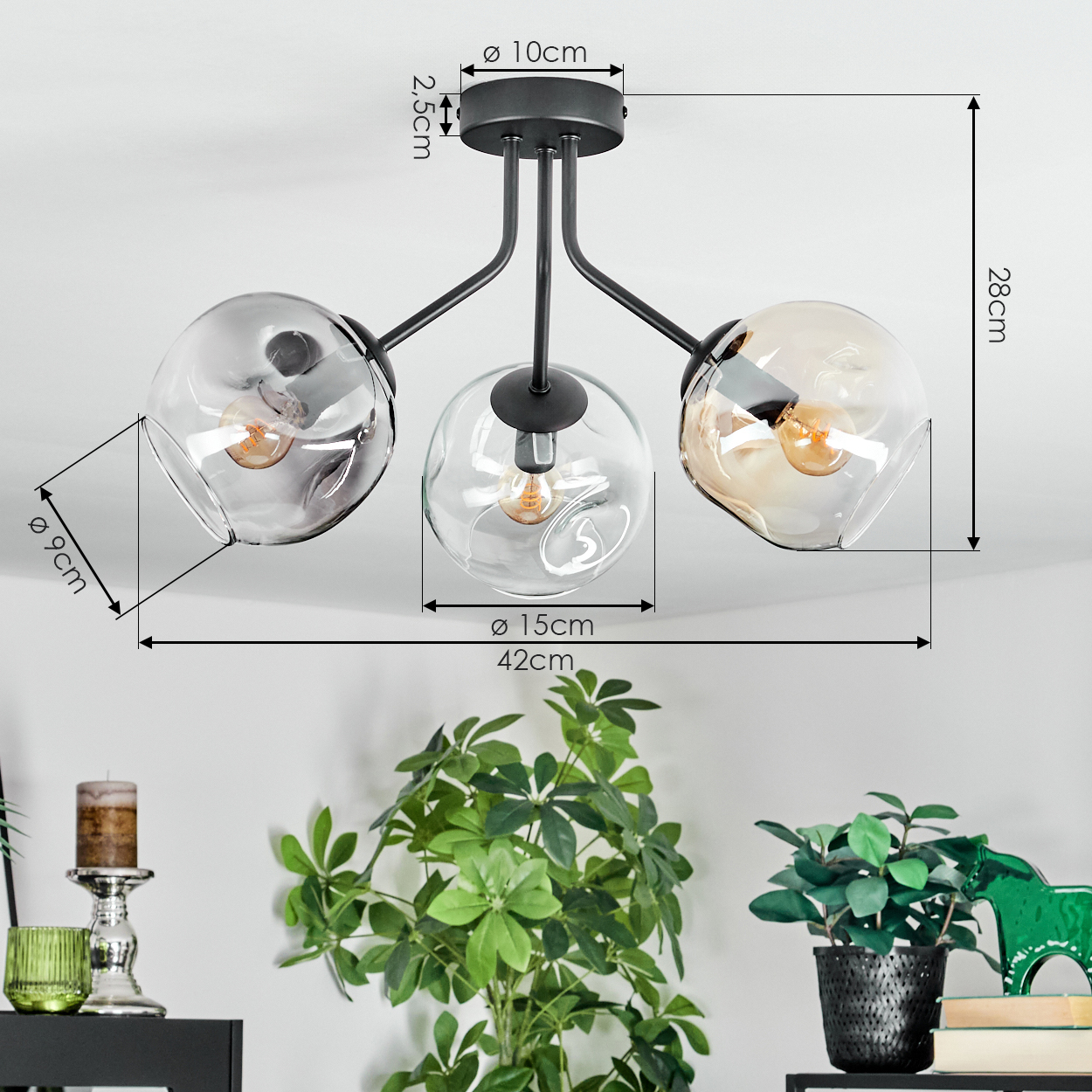 Moderne Deckenlampe mit drei Glaskugeln. Maße sind angegeben. Dekorative Pflanzen und eine Kerze im Vordergrund.