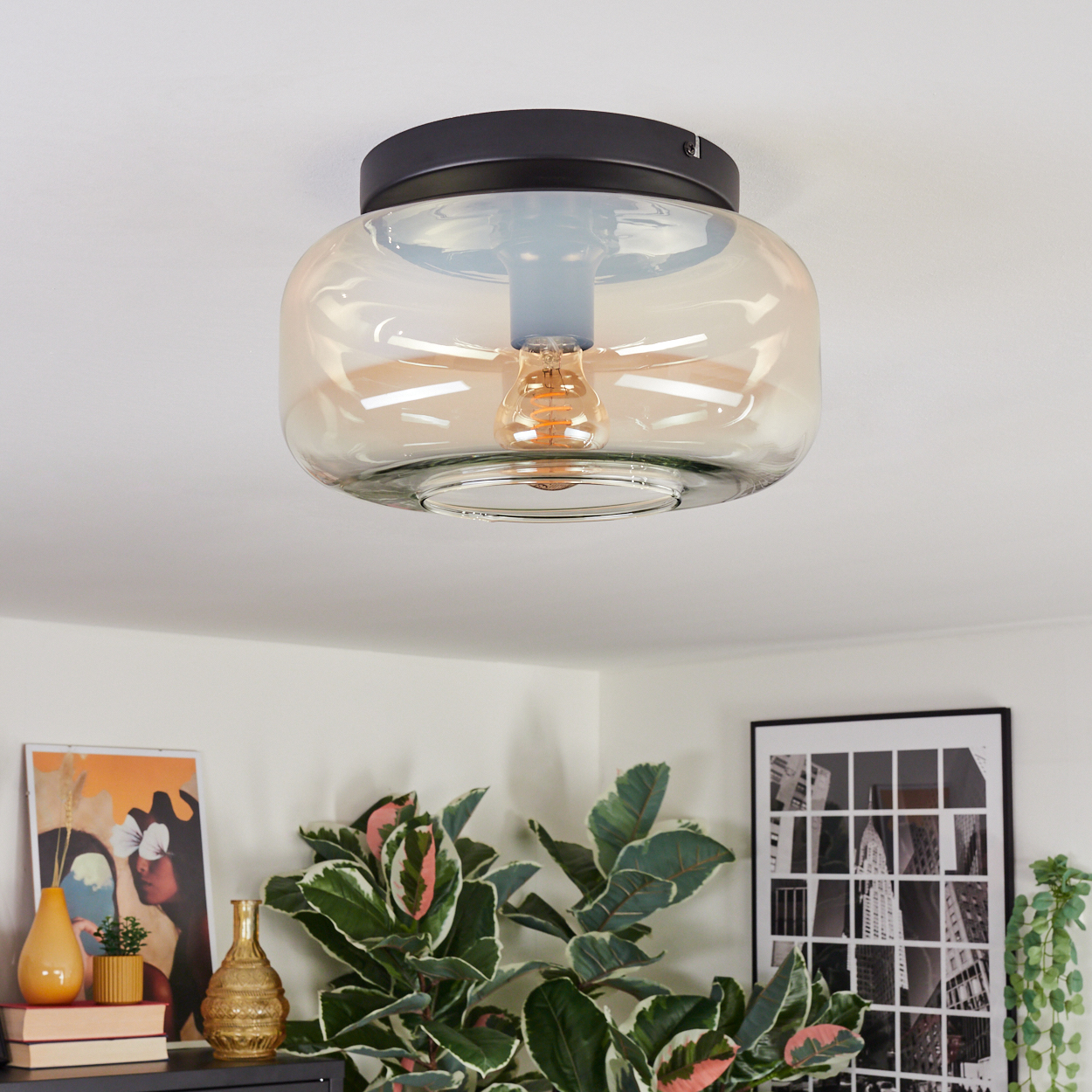 Eine Deckenlampe mit Glasschirm und sichtbarer Glühbirne. Darunter sind Pflanzen, Kunst und Vasen zu sehen, die eine gemütliche Atmosphäre schaffen.