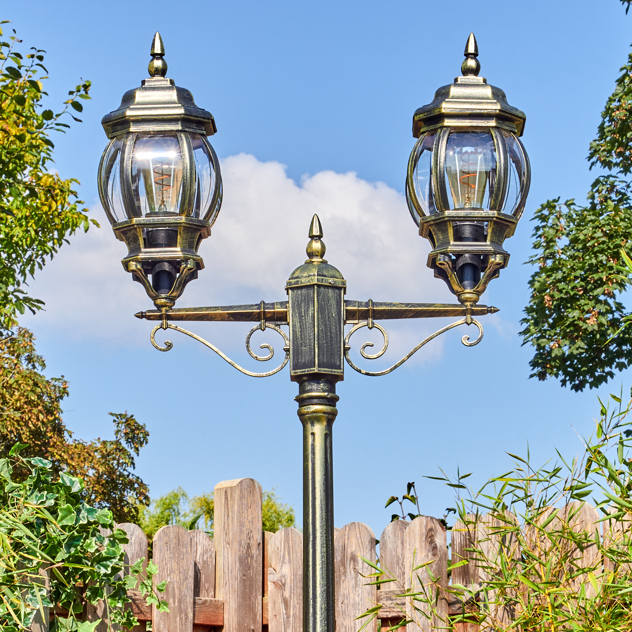 Eine dekorative Laterne mit zwei Lampen, umgeben von grünem Laub, mit einem Holzzaun im Hintergrund vor blauem Himmel.