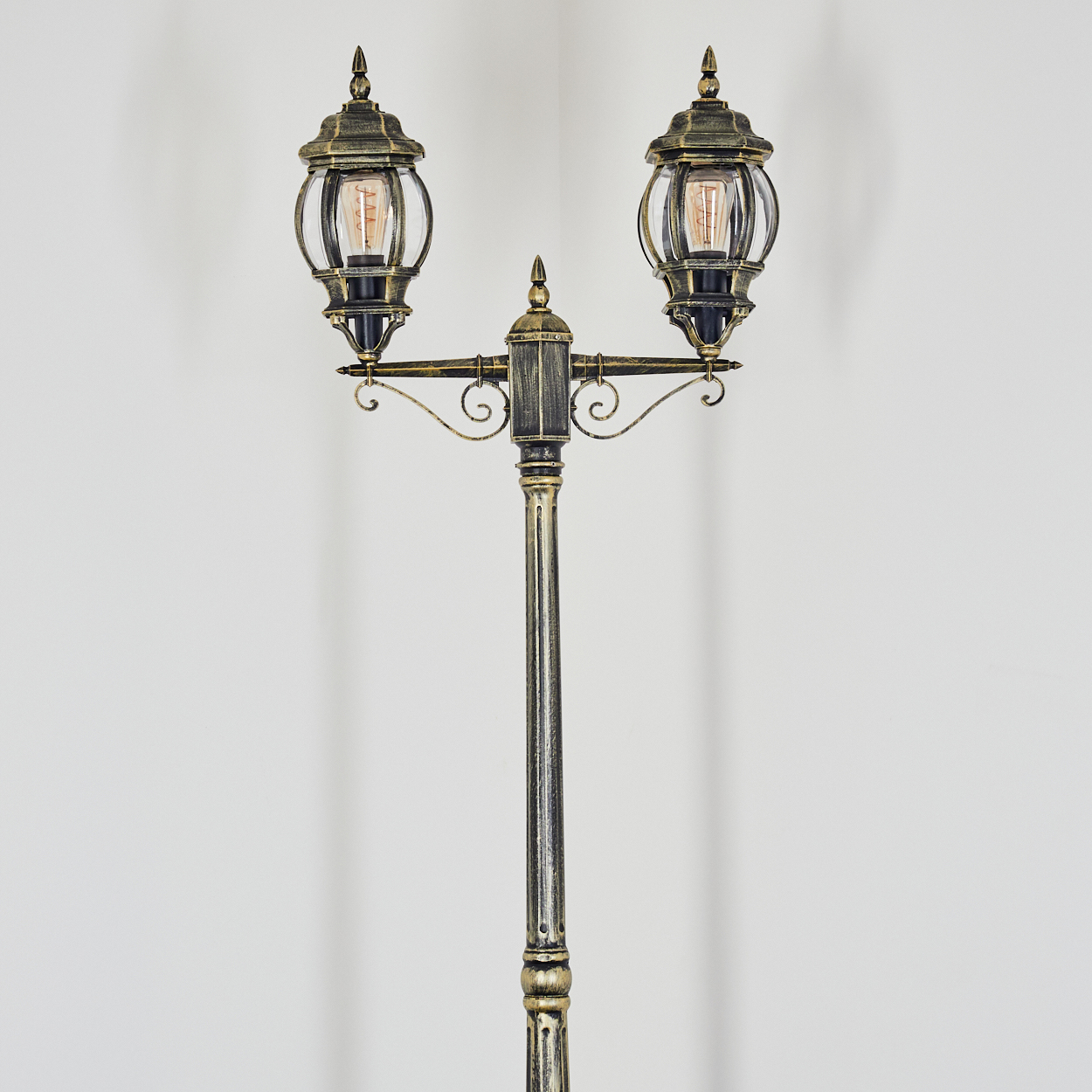 Eine verzierte Straßenlaterne mit zwei glaseingeschlossenen Lichtern. Die Lampe ist gold und schwarz vor weißem Hintergrund.