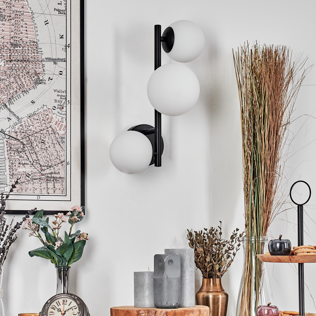 Eine moderne Wandlampe mit drei weißen Kugeln. Darunter sind eine Karte, Blumen, Kerzen und Dekorationsartikel angeordnet.