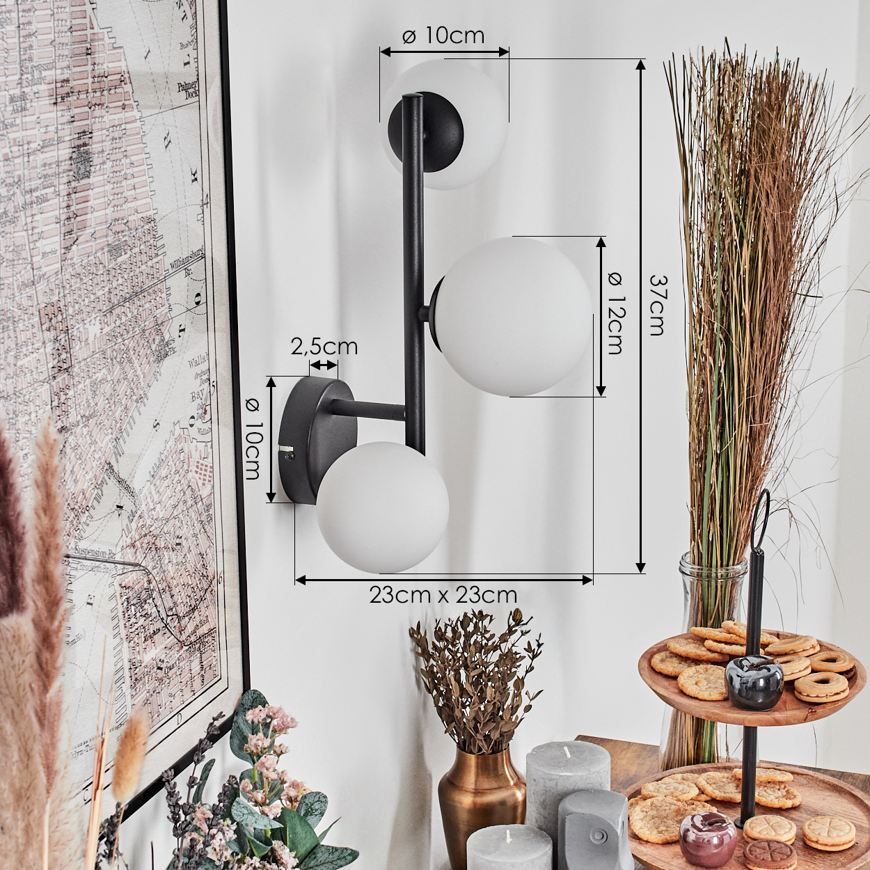 Eine moderne Wandlampe mit drei weißen Kugelleuchten. Der Hintergrund umfasst eine Karte, getrocknete Pflanzen und ein Kuchenregal.