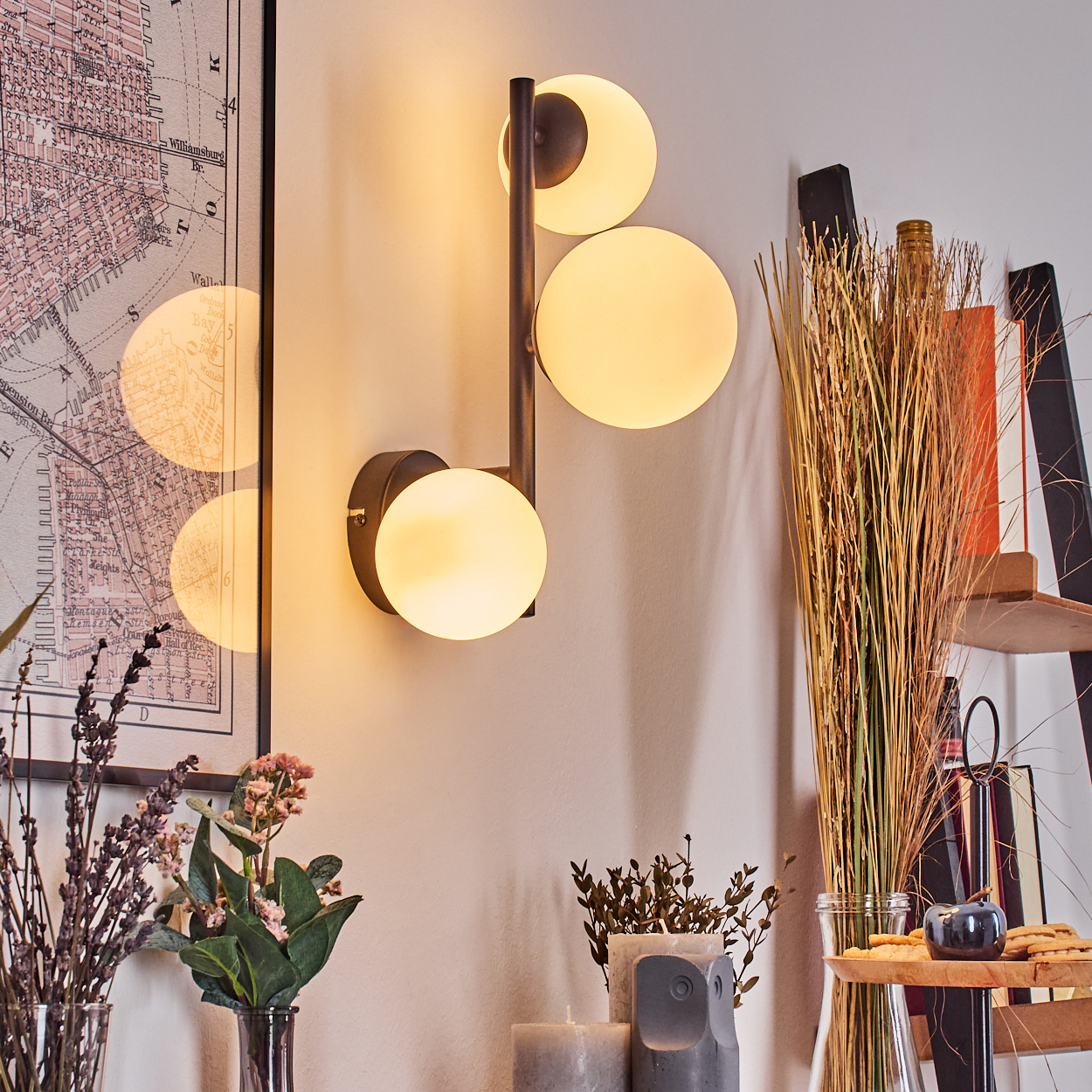 Eine moderne Wandlampe mit drei runden Lichtern. Die Wand ist mit einer Karte und Blumen verziert. Auf einem Regal stehen Kerzen und Pflanzen.