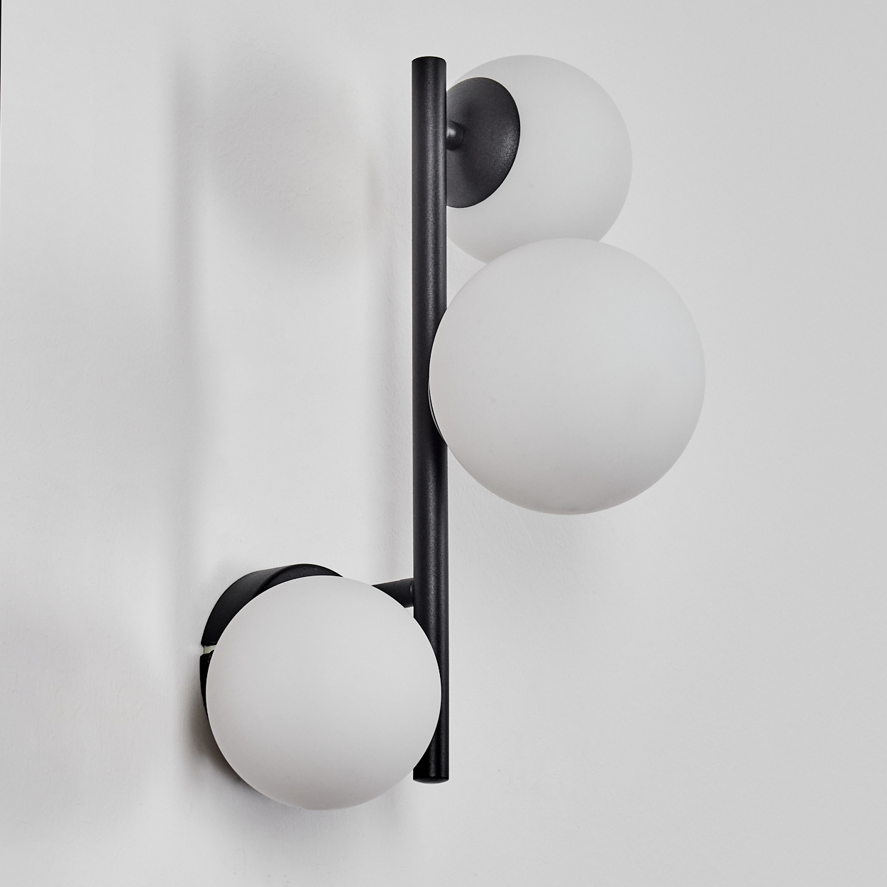 Eine moderne Wandlampe mit drei weißen Kugelleuchten und einem schwarzen Rahmen, an einer weißen Wand befestigt.