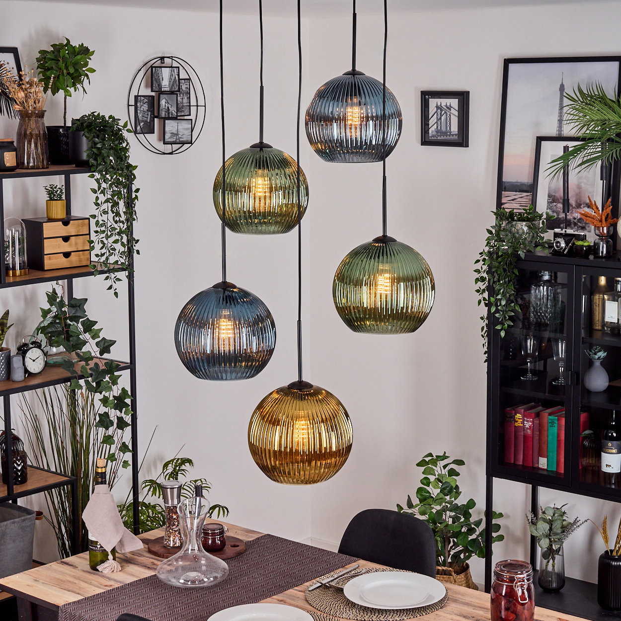 Esszimmer mit Hängelampen. Tisch mit Tellern, einer Karaffe und Pflanzen. Regale mit Dekor und ein schwarzer Schrank.