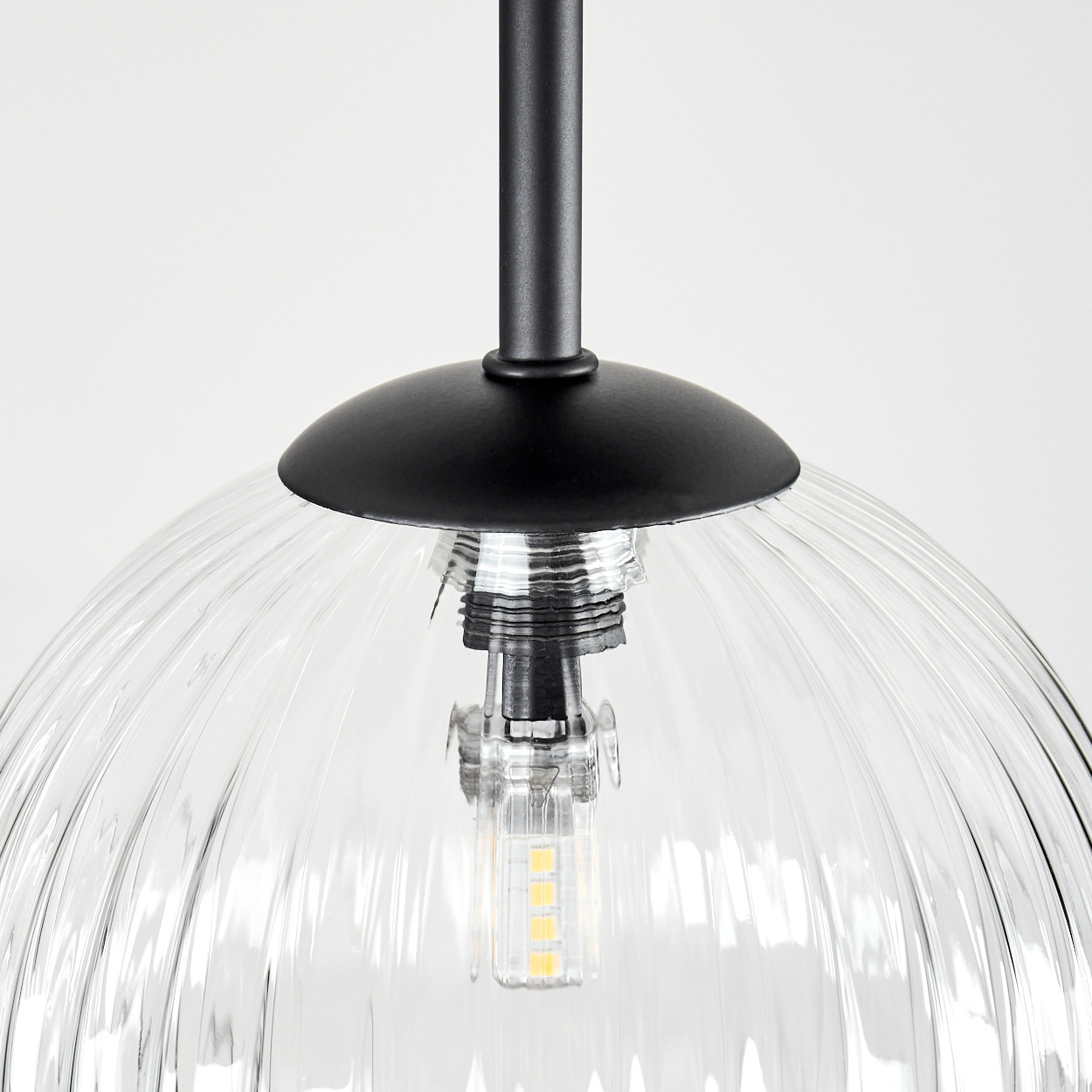 Nahaufnahme einer Pendelleuchte. Sie hat einen schwarzen Deckel, einen transparenten, gerippten Glasschirm und eine sichtbare LED-Glühbirne.