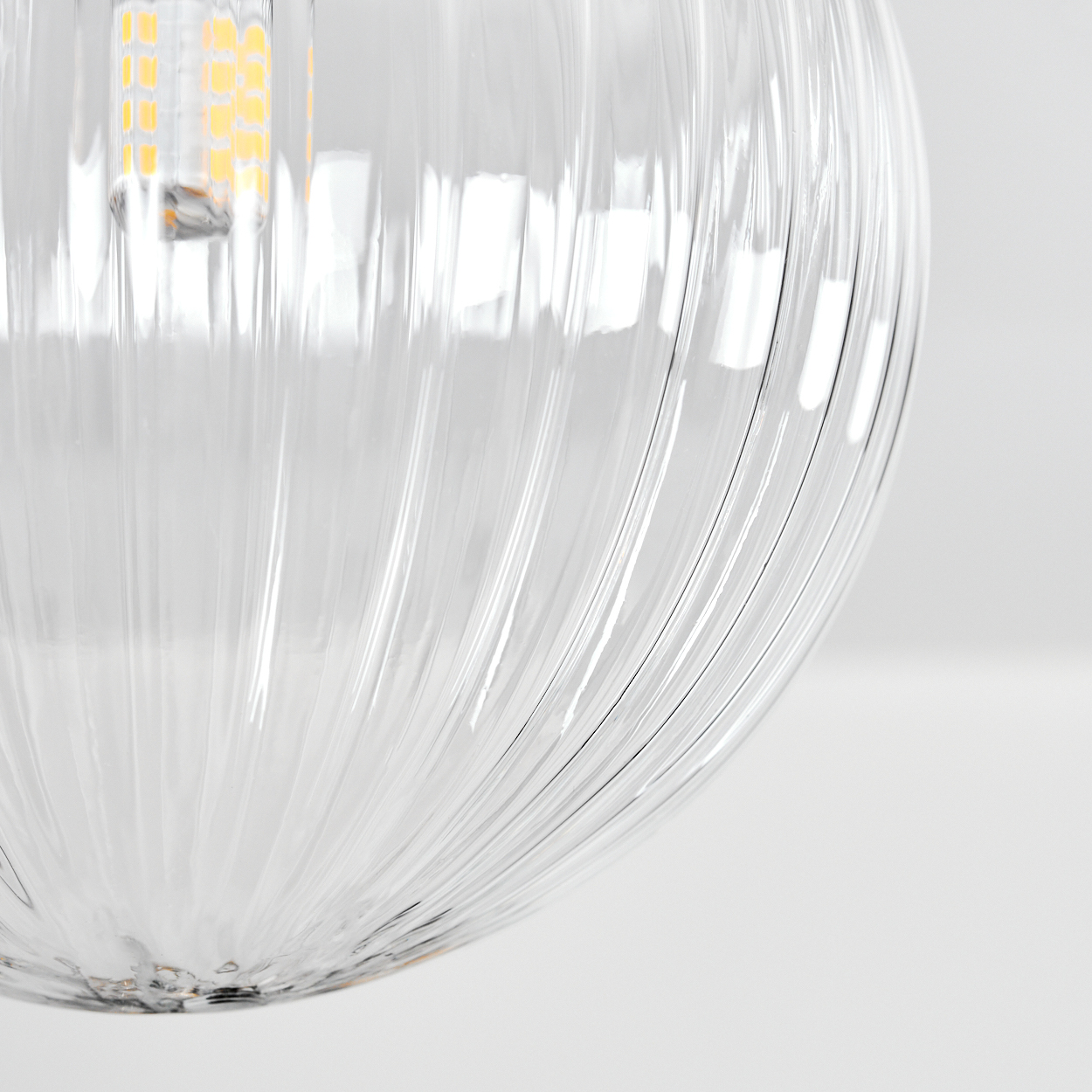 Eine klare, gerippte Glas-Pendelleuchte mit einer leuchtenden LED-Glühbirne im Inneren. Der Hintergrund ist eine saubere, weiße Oberfläche.