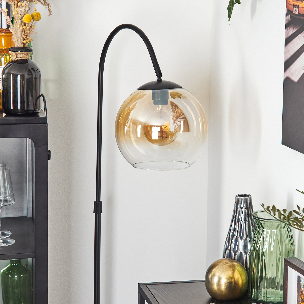 Eine moderne Stehlampe mit bernsteinfarbenem Glasschirm. Die Szene umfasst dekorative Vasen und eine goldene Kugel auf einem dunklen Tisch.