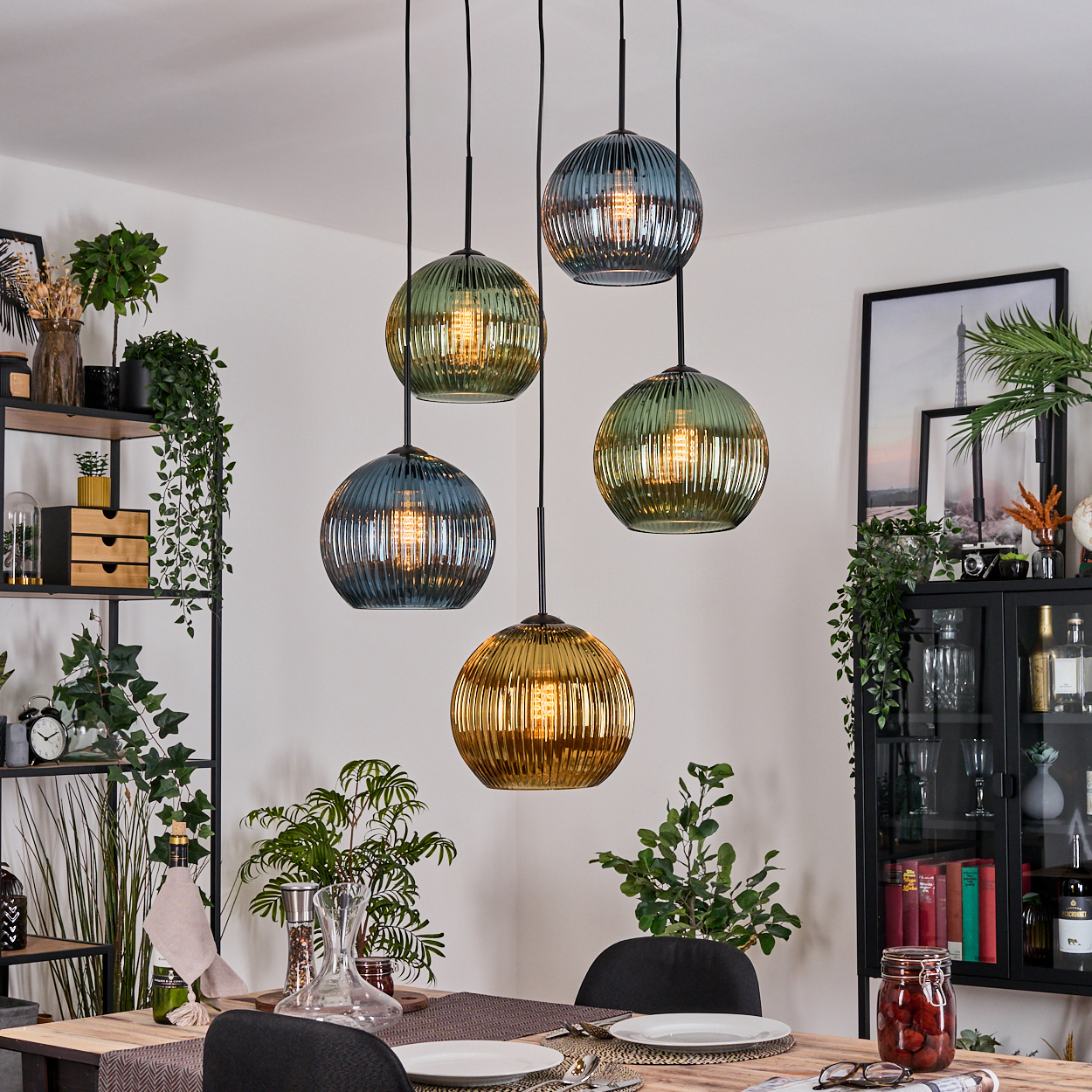 Esszimmer mit Hängelampen. Lampen sind Glaskugeln in verschiedenen Farben. Tisch mit Tellern und Stühlen. Regale und Pflanzen im Hintergrund.