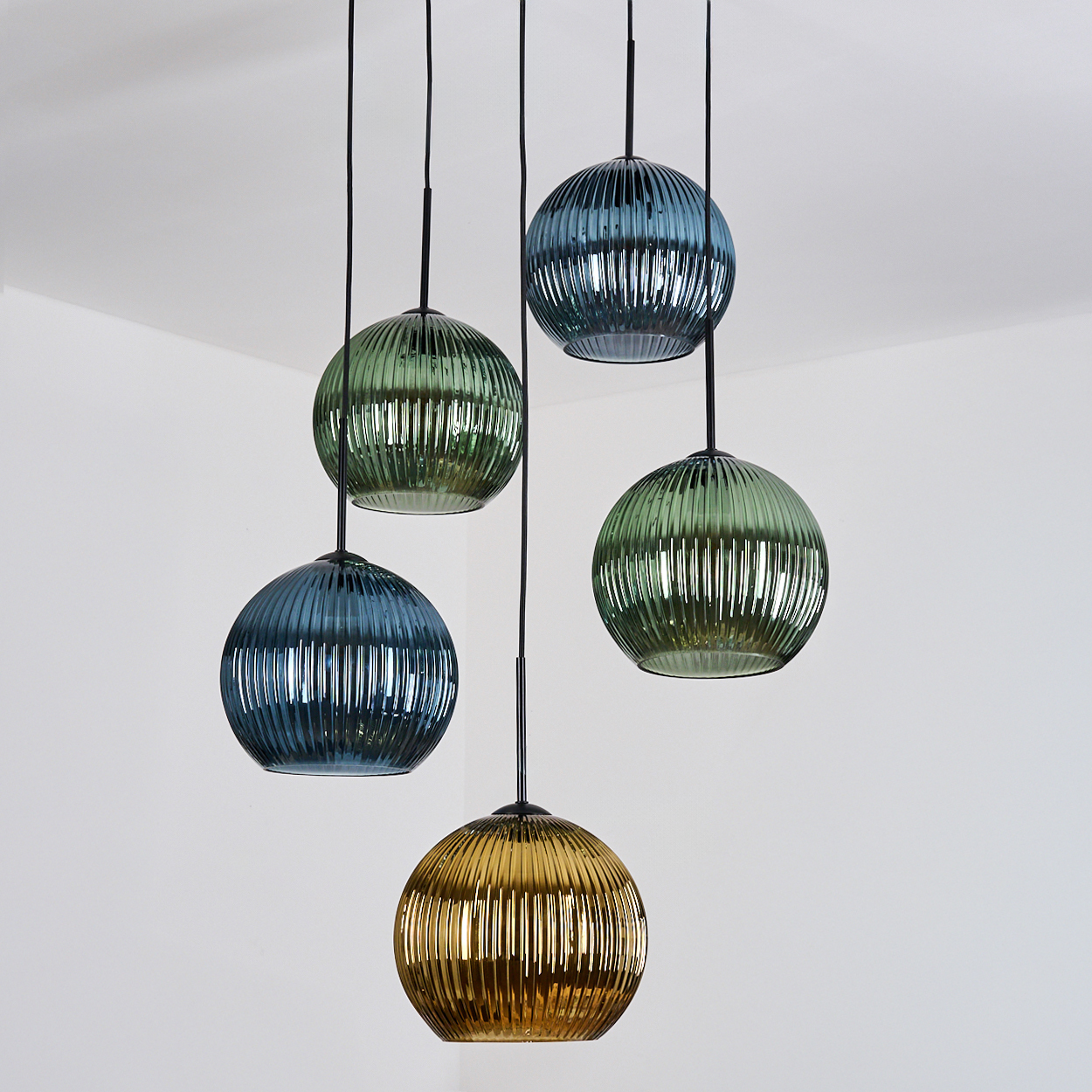 Fünf hängende Pendelleuchten aus geripptem Glas. Farben sind Blau, Grün und Gold vor einer weißen Wand.