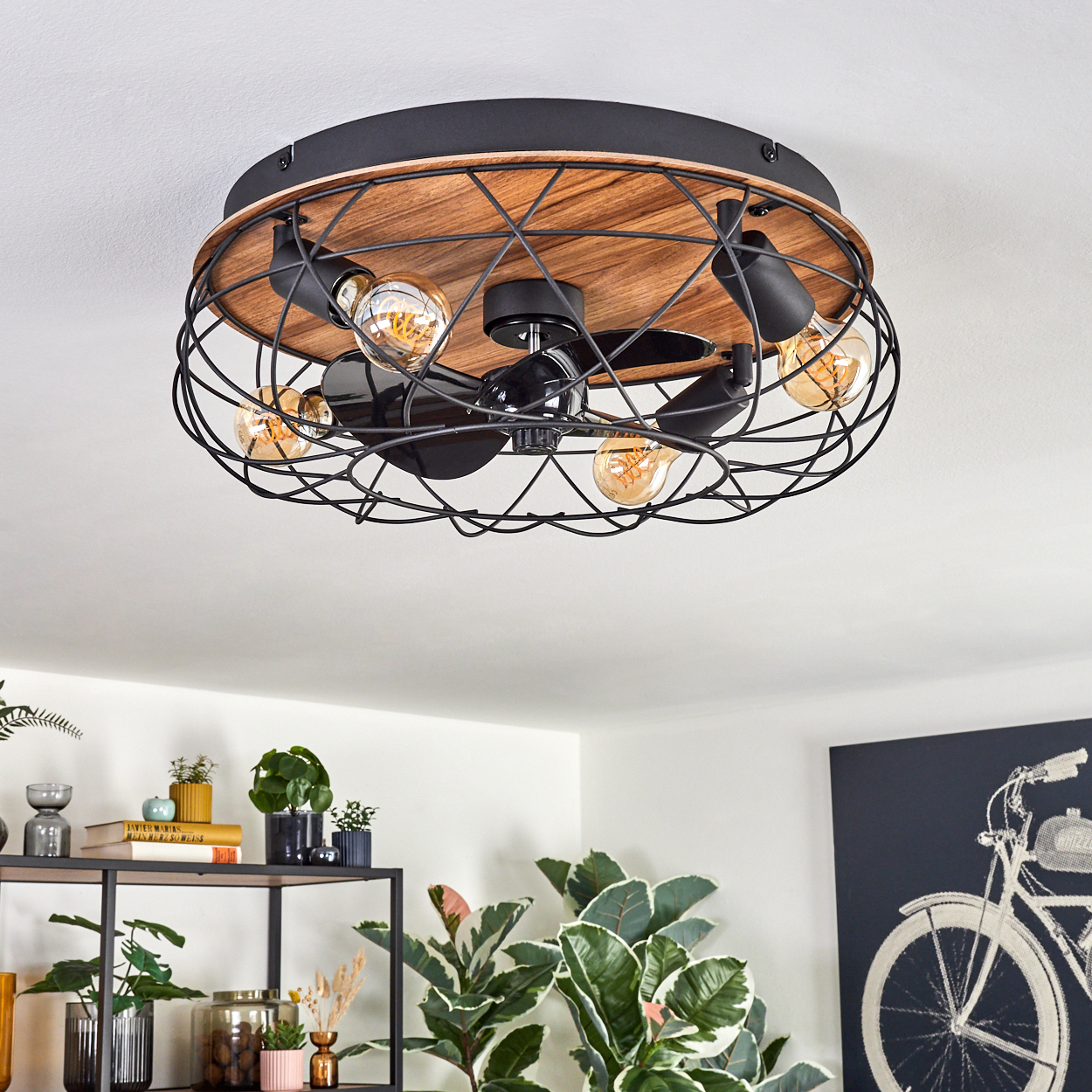 Deckenventilator mit freiliegenden Glühbirnen und Holzsockel. Darunter sind Pflanzen und ein Fahrradkunstwerk zu sehen.