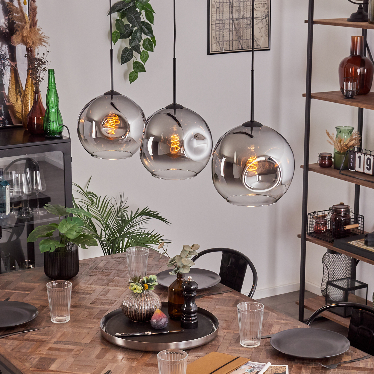 Esszimmer mit gedecktem Tisch. Drei Glaskugel-Pendelleuchten hängen darüber. Regale und Pflanzen schmücken den Raum.