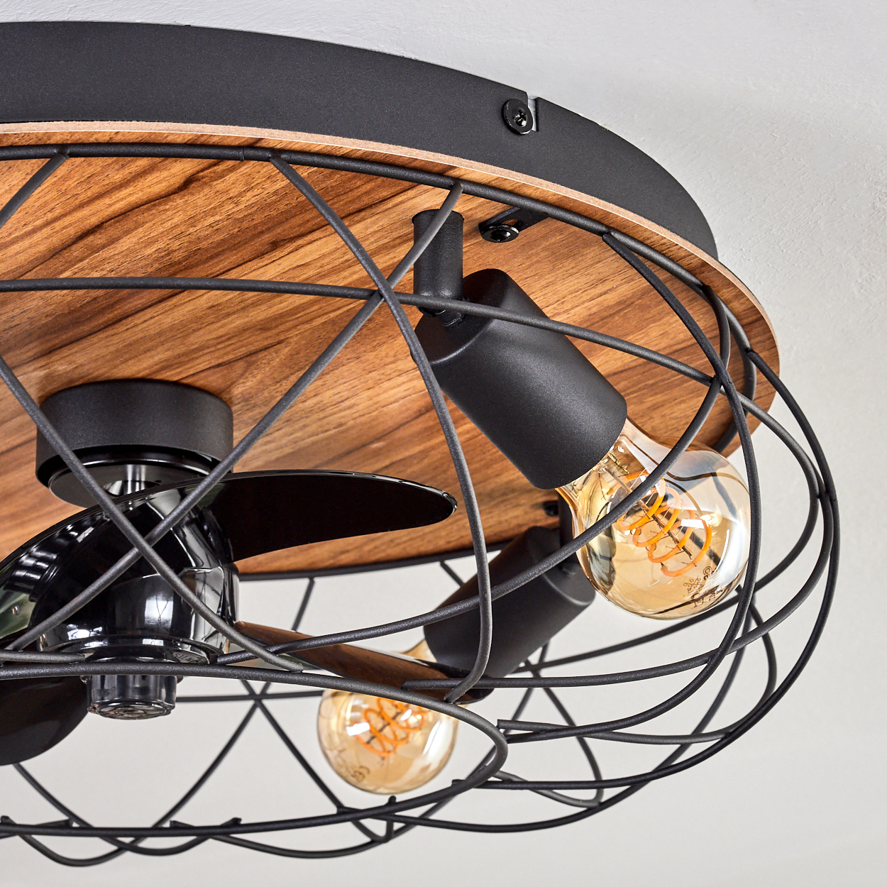Ein Deckenventilator mit freiliegenden Glühbirnen. Er hat einen schwarzen Metallkäfig, eine Holzbasis und schwarze Lüfterblätter. Die Glühbirnen sind beleuchtet.
