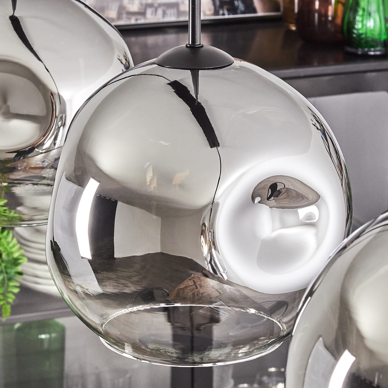 Drei verspiegelte Pendelleuchten aus Glas mit schwarzen Halterungen. Sie reflektieren die Umgebung und das Licht. Der Hintergrund zeigt Regale und Pflanzen.