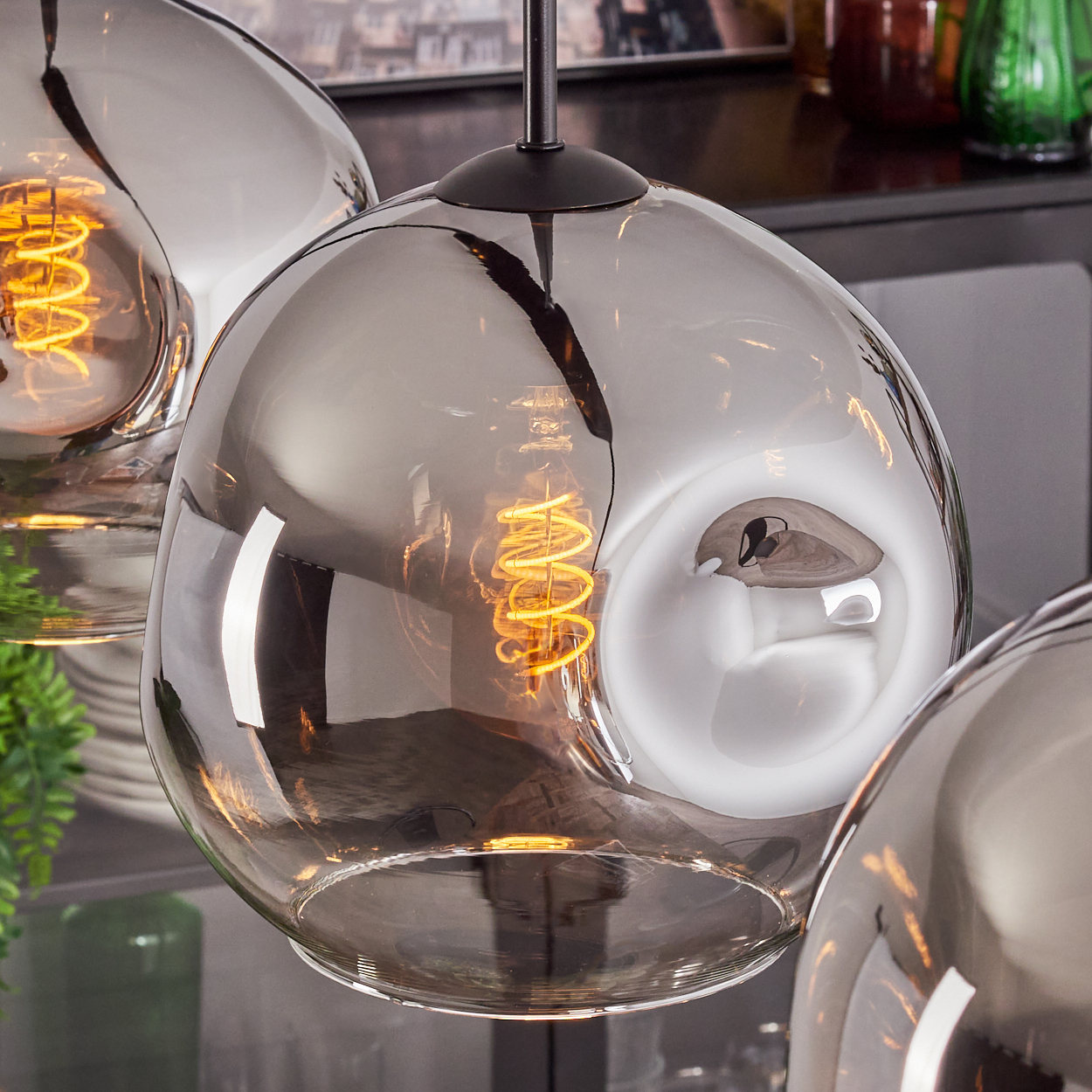 Drei rauchfarbene Glas-Pendelleuchten mit warmen Wendelglühbirnen. Reflektierende Oberflächen zeigen die Umgebung. Schwarze Halterung.