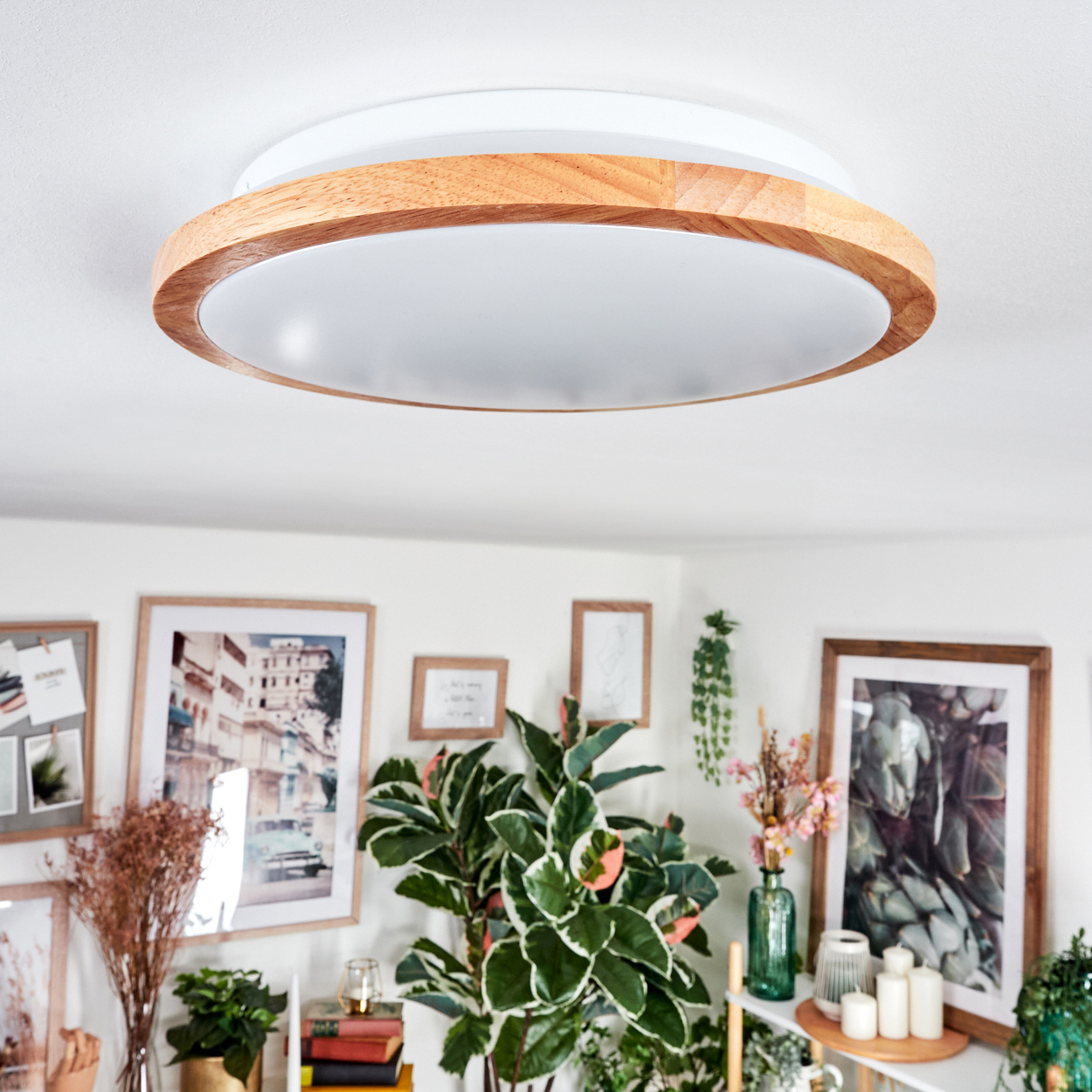 Eine moderne Deckenlampe mit Holzrahmen ist zentriert. Darunter ist ein Raum mit Pflanzen, Bildern und Kerzen zu sehen.