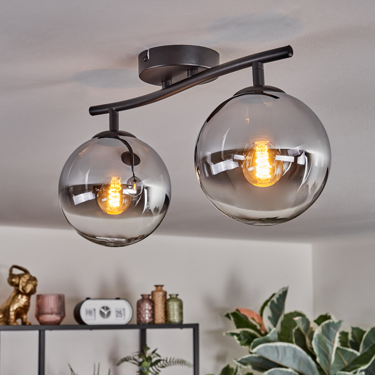 Eine moderne Deckenleuchte mit zwei kugelförmigen Glasschirmen, jede mit einer warm getönten Glühbirne. Ein Regal darunter hält Dekoration.
