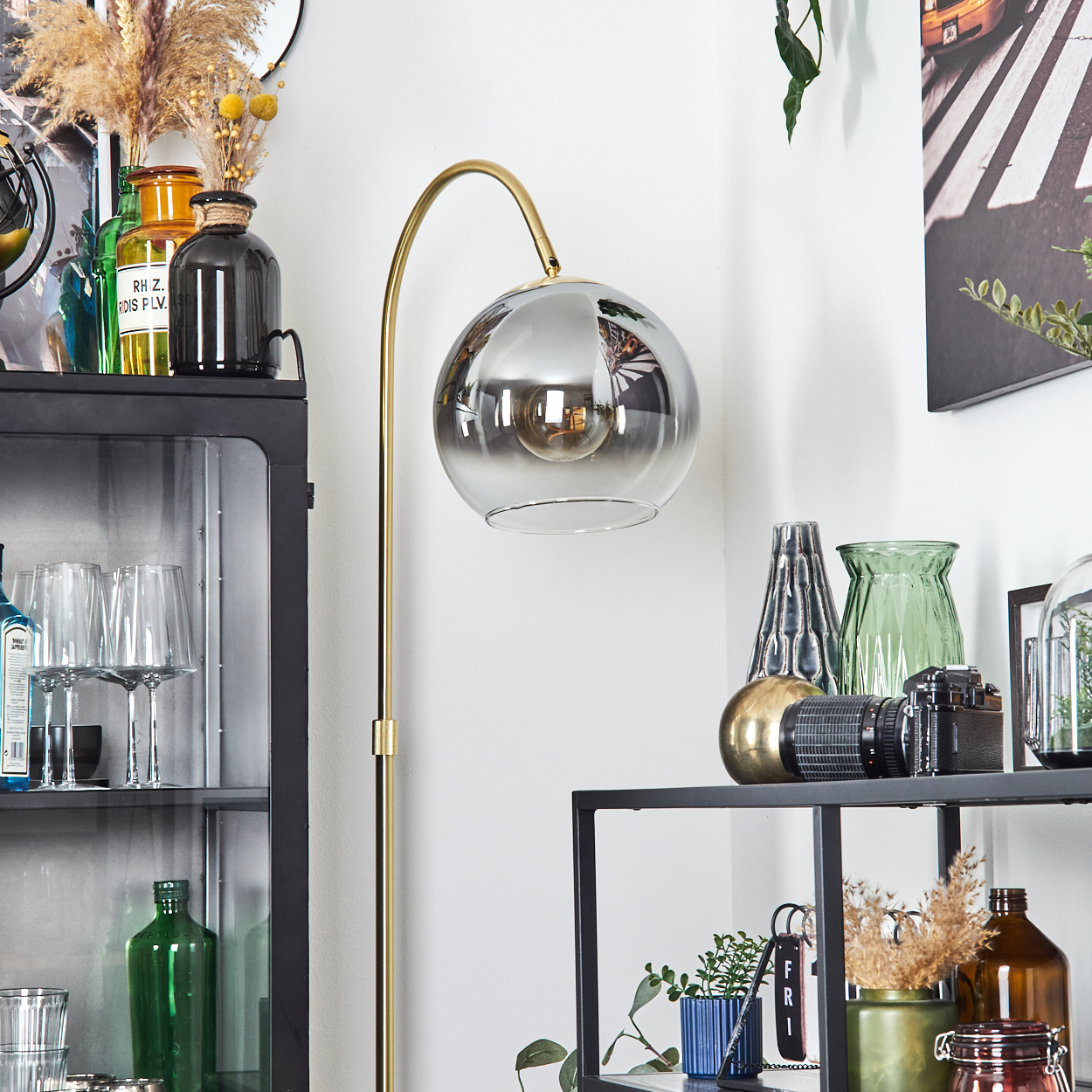 Modernes Interieur mit goldener Lampe, Glasregalen mit verschiedenen Objekten und Wandkunst mit Straßenszene.