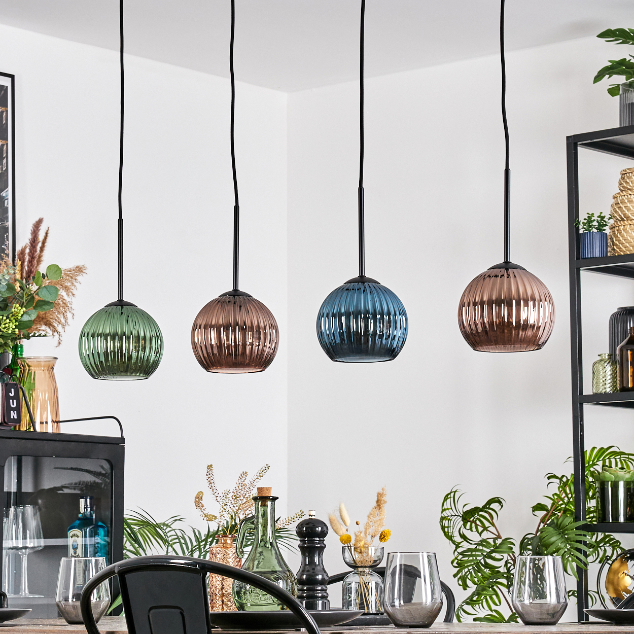 Vier bunte Glas-Pendelleuchten hängen über einem Esstisch mit Gläsern, Pflanzen und anderen Dekorationen.