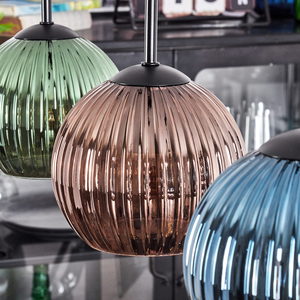 Drei Pendelleuchten mit gerippten Glasschirmen in Grün, Bronze und Blau. Schwarze Leuchten hängen von der Decke.
