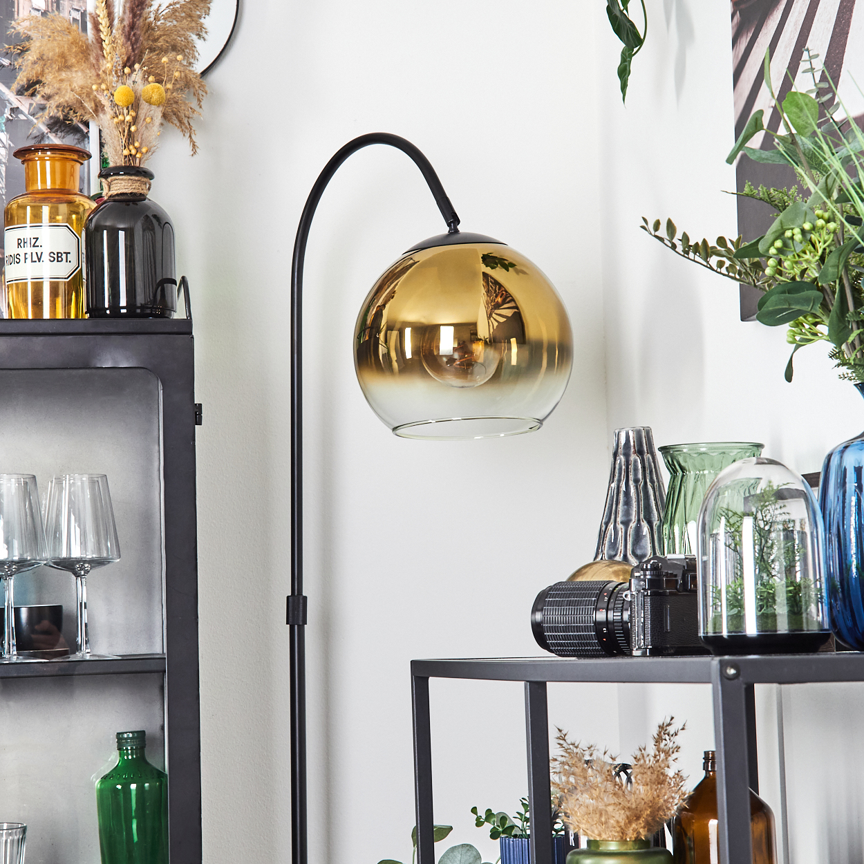 Eine goldene Lampe, Regale mit Vasen, eine Kamera und Gläser sind sichtbar. Der Hintergrund ist weiß.